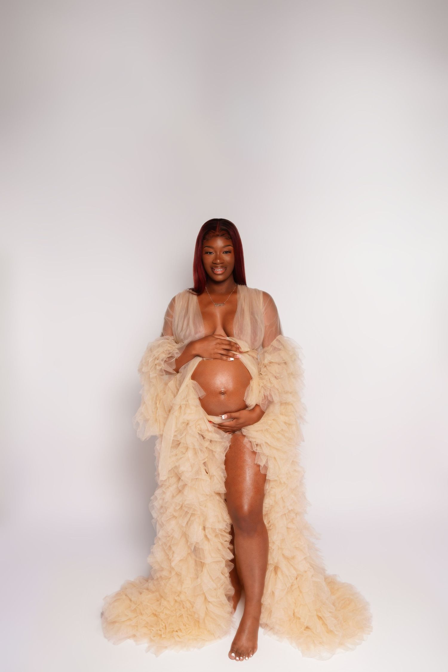 Pregnant woman in sheer beige ruffled maternity gown cradling baby bump, smiling against white studio background.