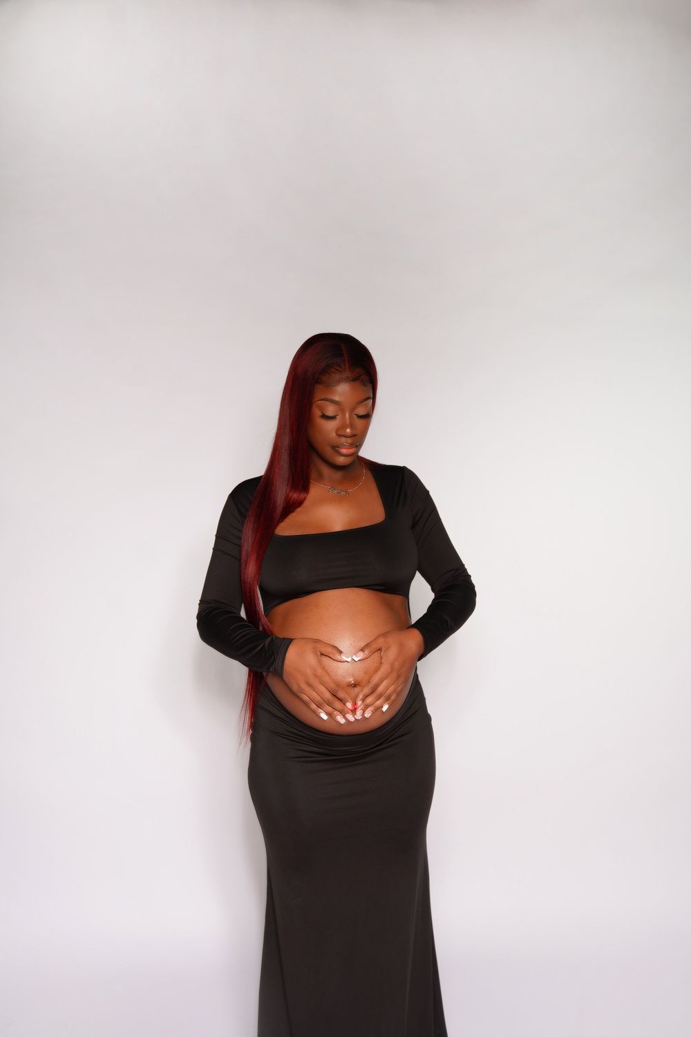 Pregnant Black woman with red hair in black crop top and skirt cradling baby bump against white background.