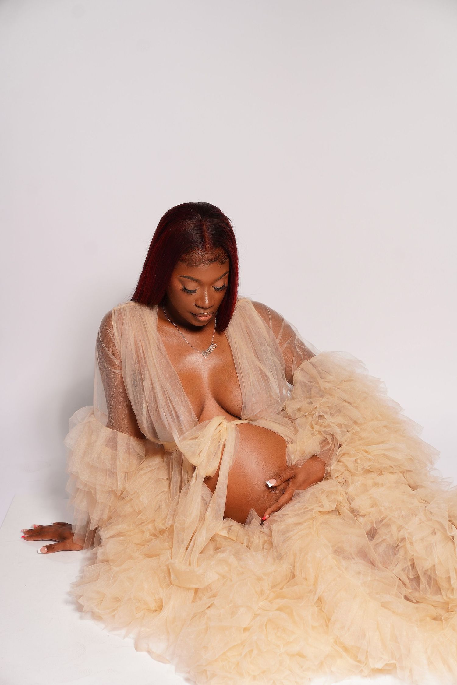 Expectant mother in tulle champagne robe sits gazing down at baby bump in studio.