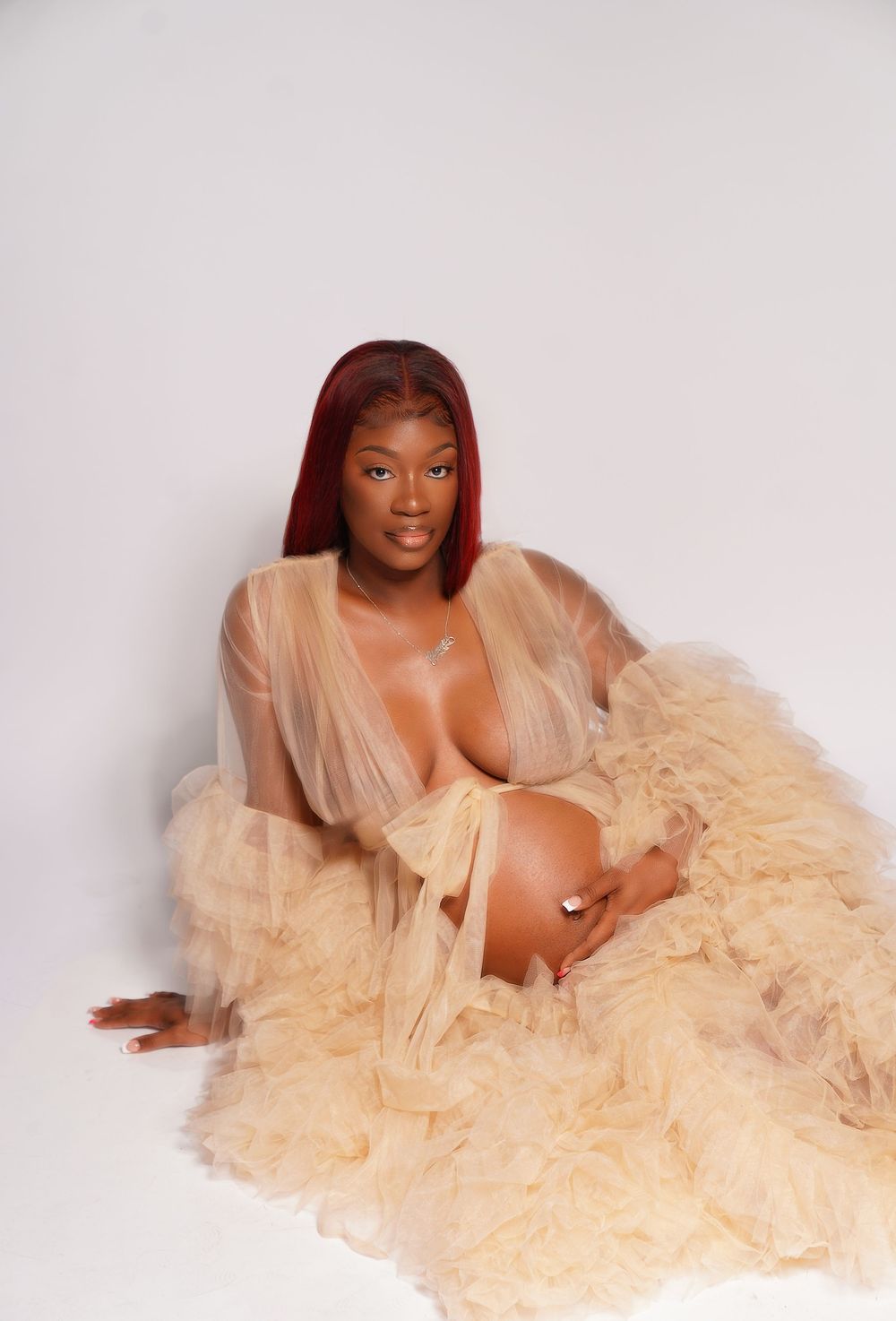 Pregnant woman in sheer champagne ruffle robe poses gracefully on white studio background.