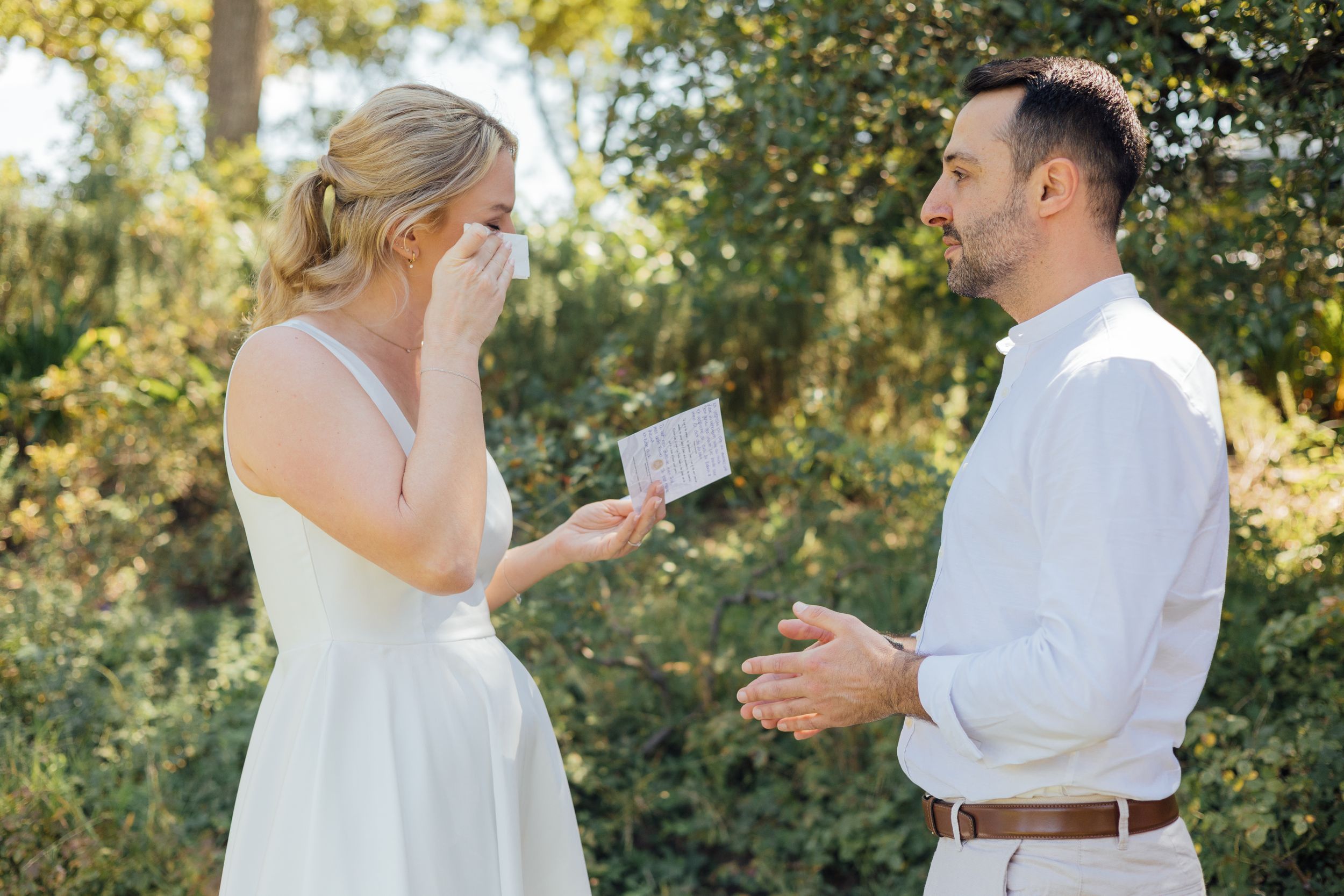 Emotional bride reads a card while groom smiles outdoors in wedding attire surrounded by lush greenery.