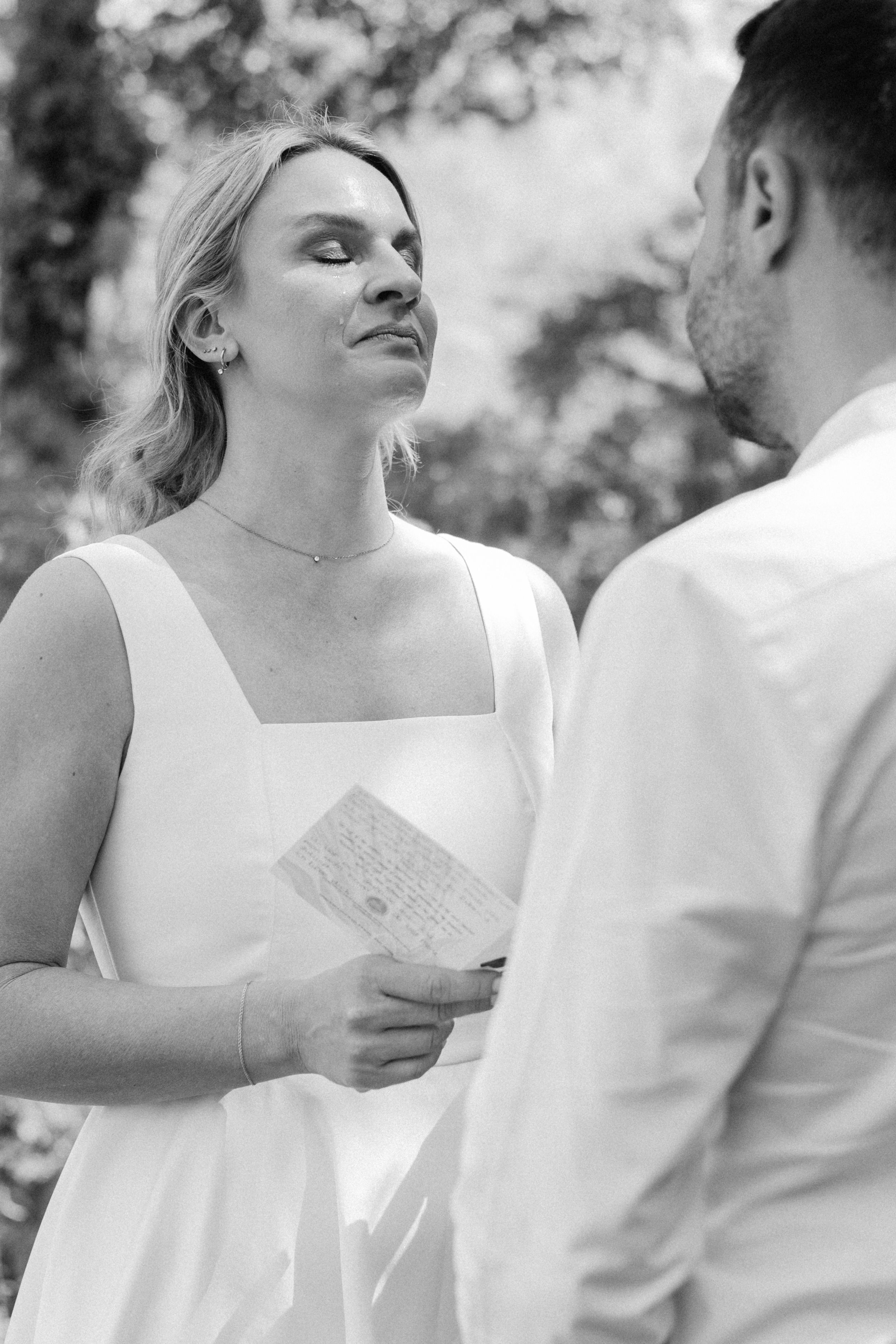Black and white photo of a bride in white dress reading vows to groom outdoors at wedding ceremony.
