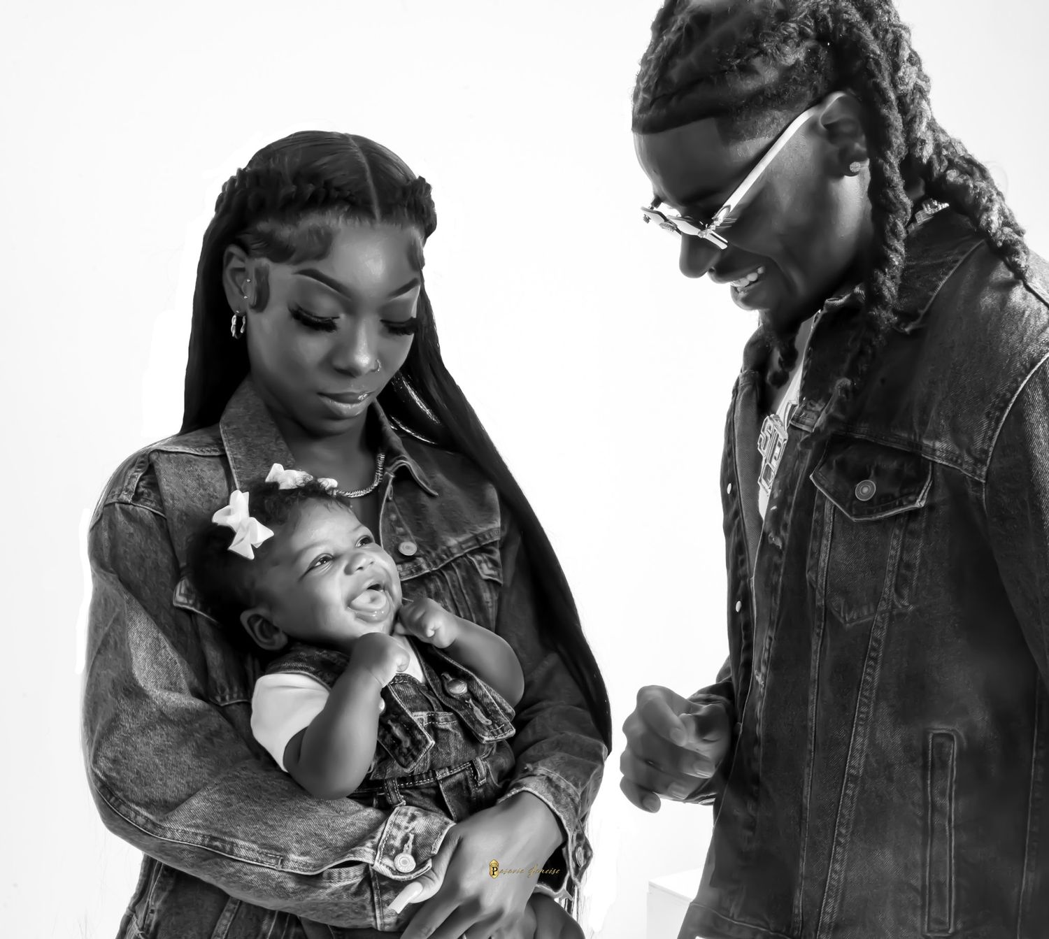 Black and white photo of a couple in denim jackets smiling at their happy baby girl with bow hair clips.