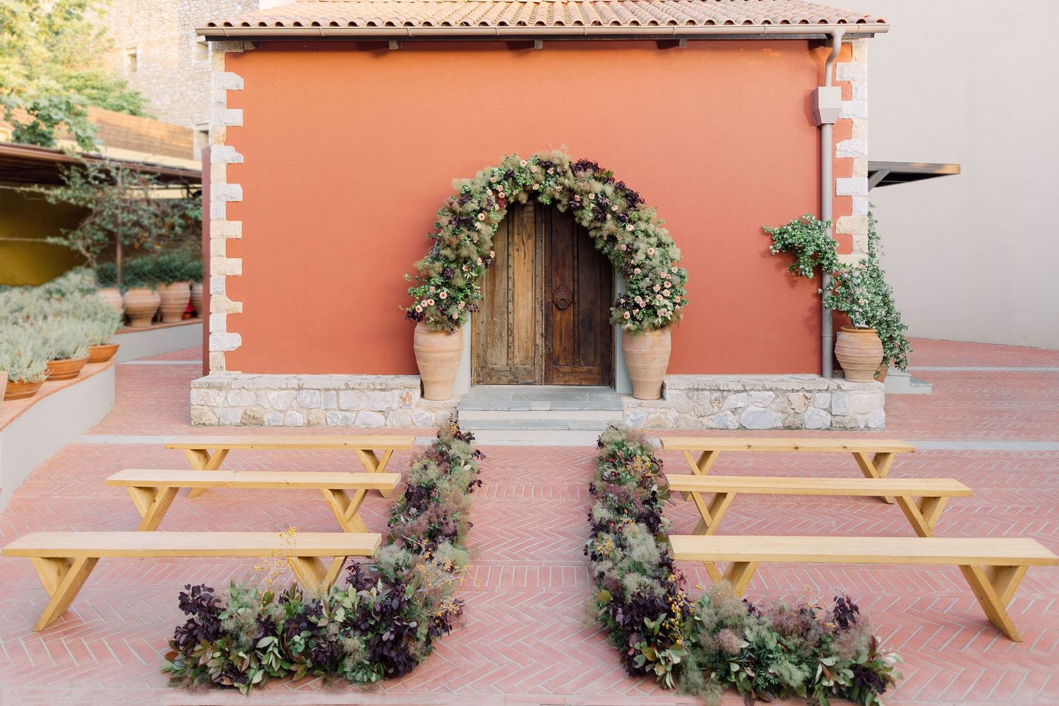 Rustic wooden picnic tables arranged in front of a salmon-colored wall with decorative greenery and archways.