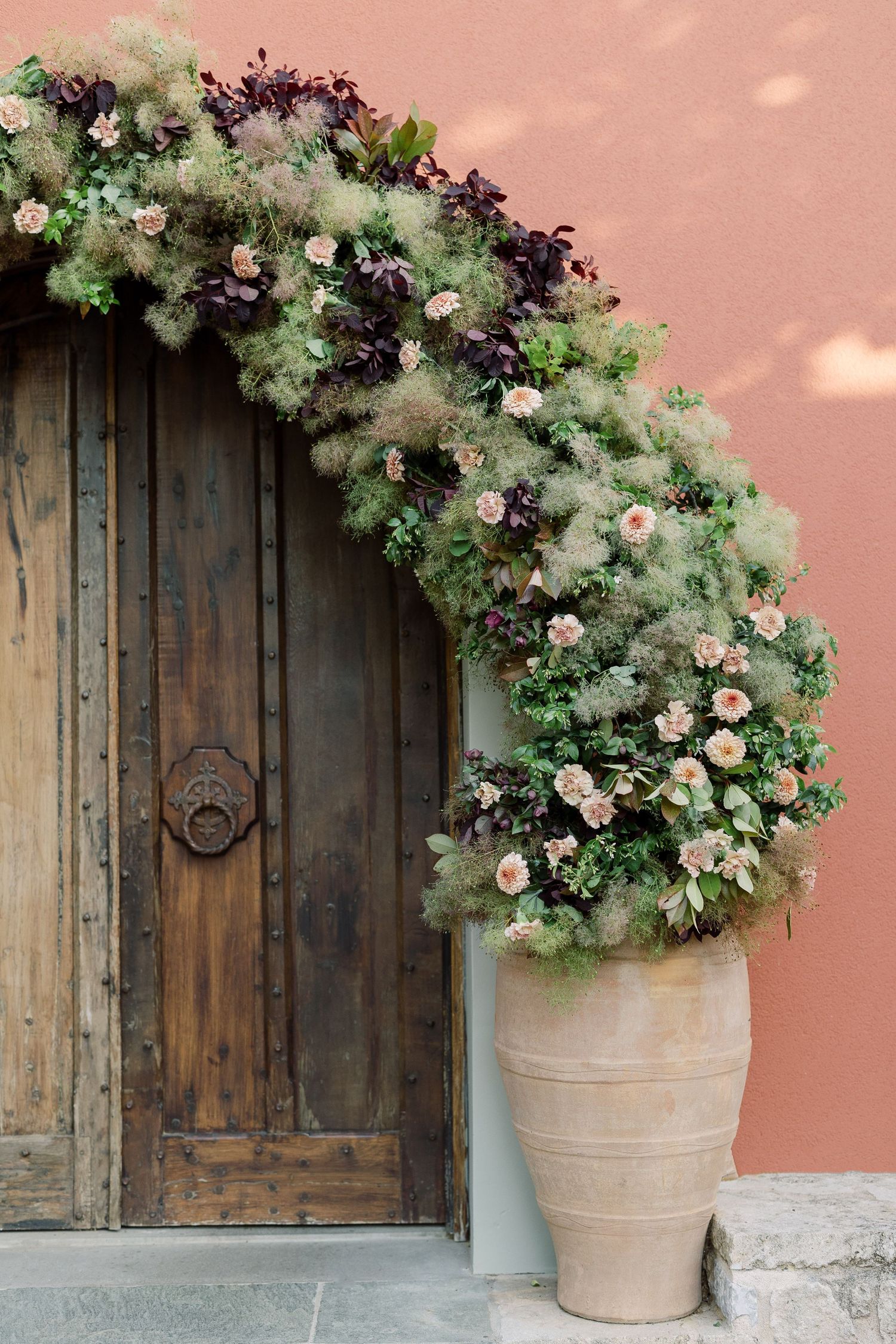 Lush floral arrangement with pink roses and greenery cascades over a rustic wooden door beside a terracotta pot.