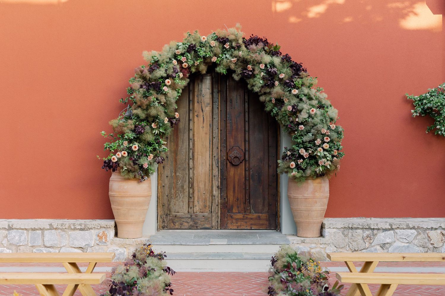 Wooden door framed by lush floral arrangements against a terracotta wall with outdoor seating.