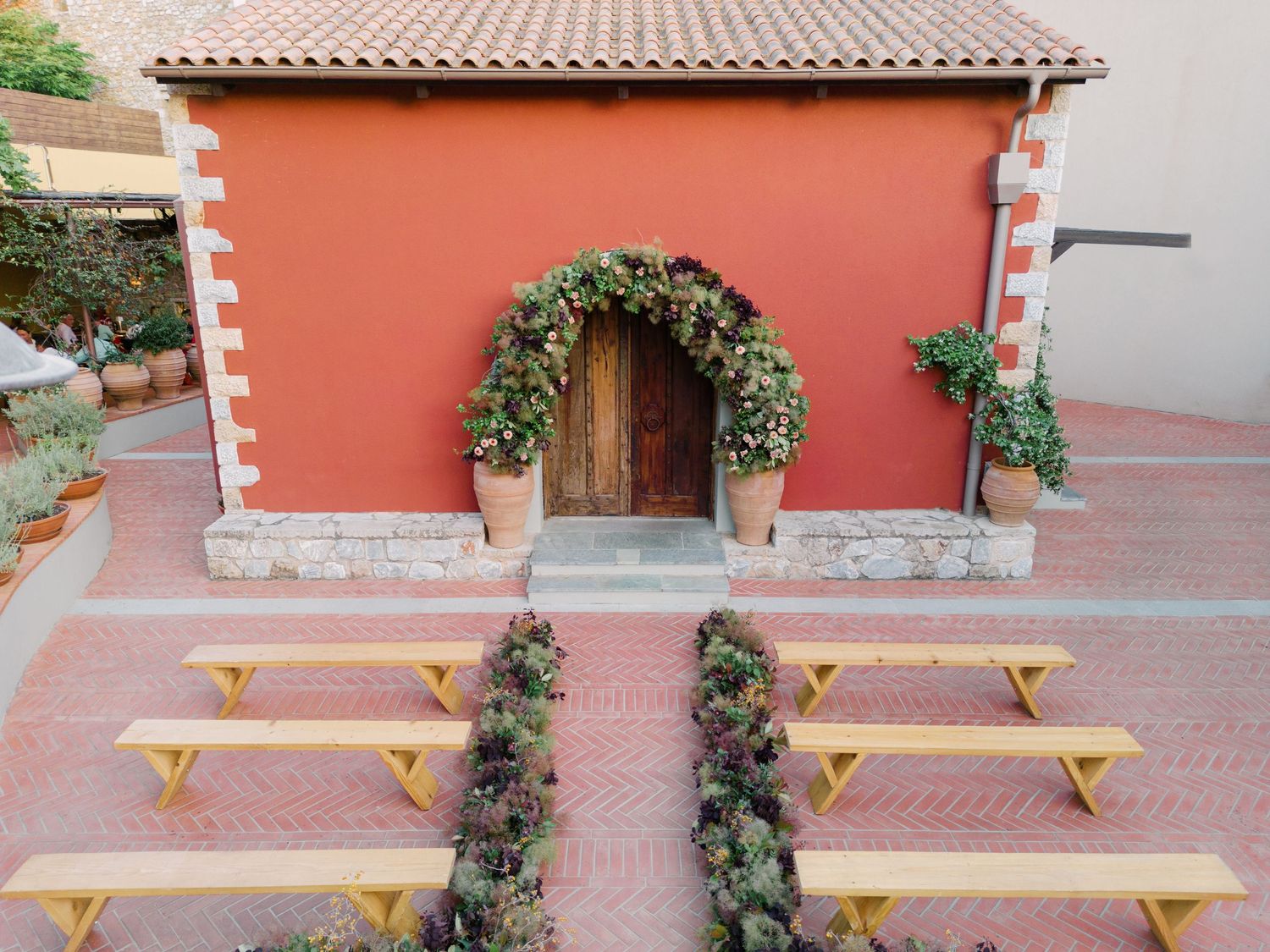 Small coral-colored building with wooden door adorned by greenery arch and yellow benches arranged in rows on patio.
