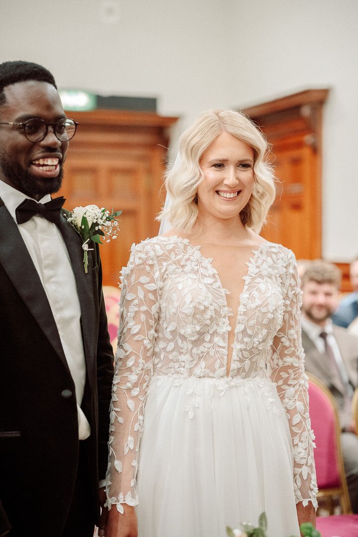 A happy couple at their wedding ceremony, with the bride wearing a long-sleeved lace gown and the groom in a black tuxedo.