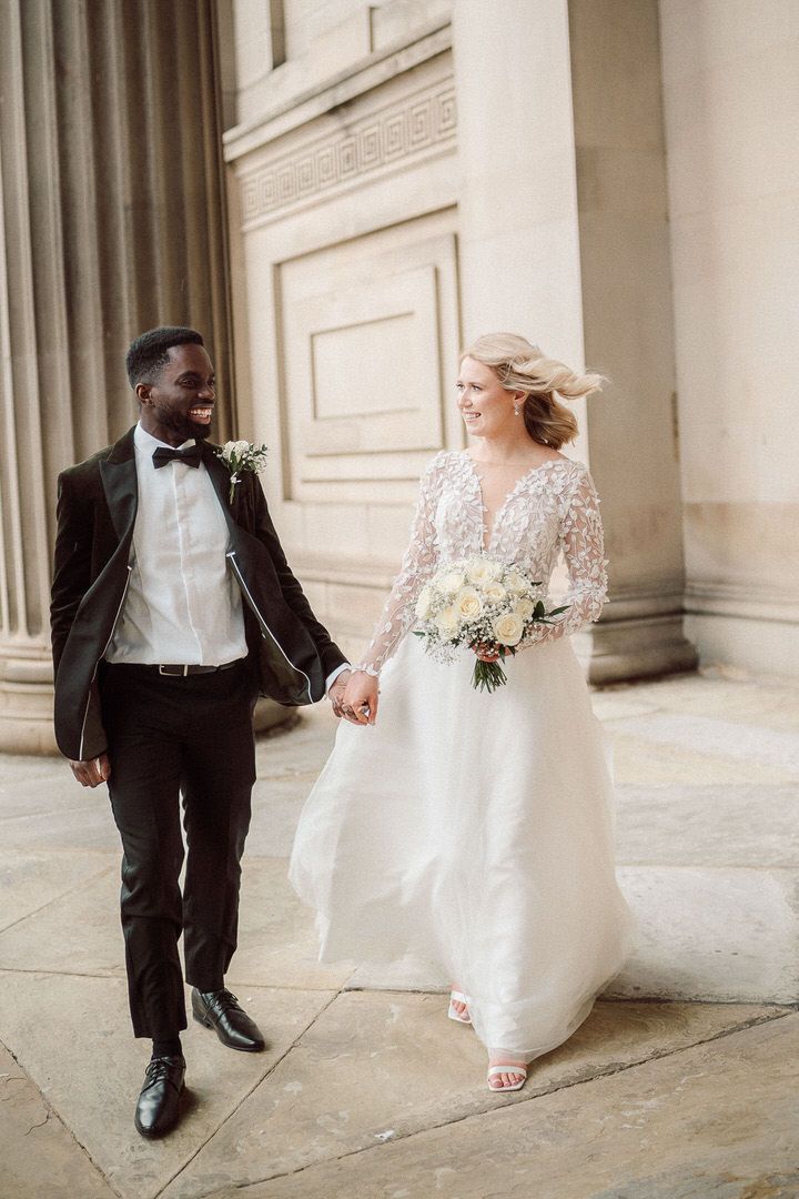 A joyful couple walking together on their wedding day outside a grand building with classical columns.