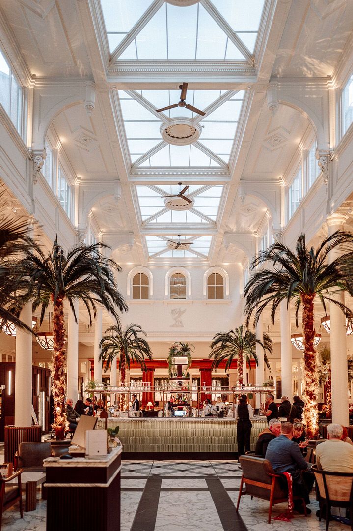 Elegant hotel atrium with glass ceiling, ornate chandeliers and palm trees surrounding a central seating area.