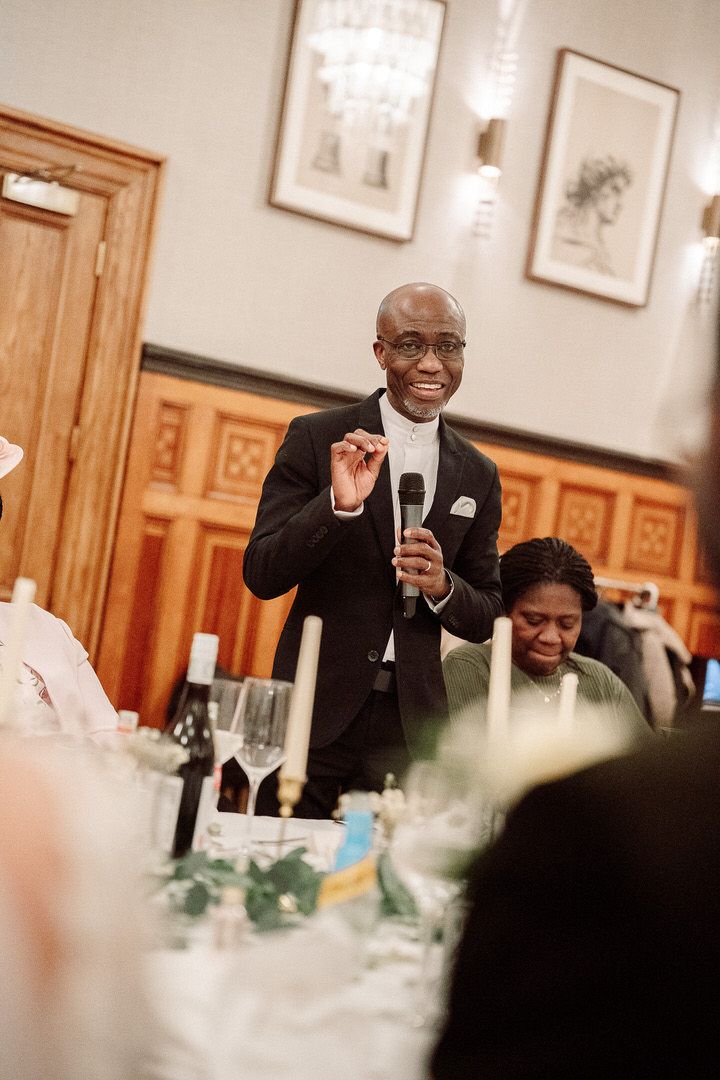 A speaker in a black suit delivers a speech while standing at a formal dinner reception with table settings visible.