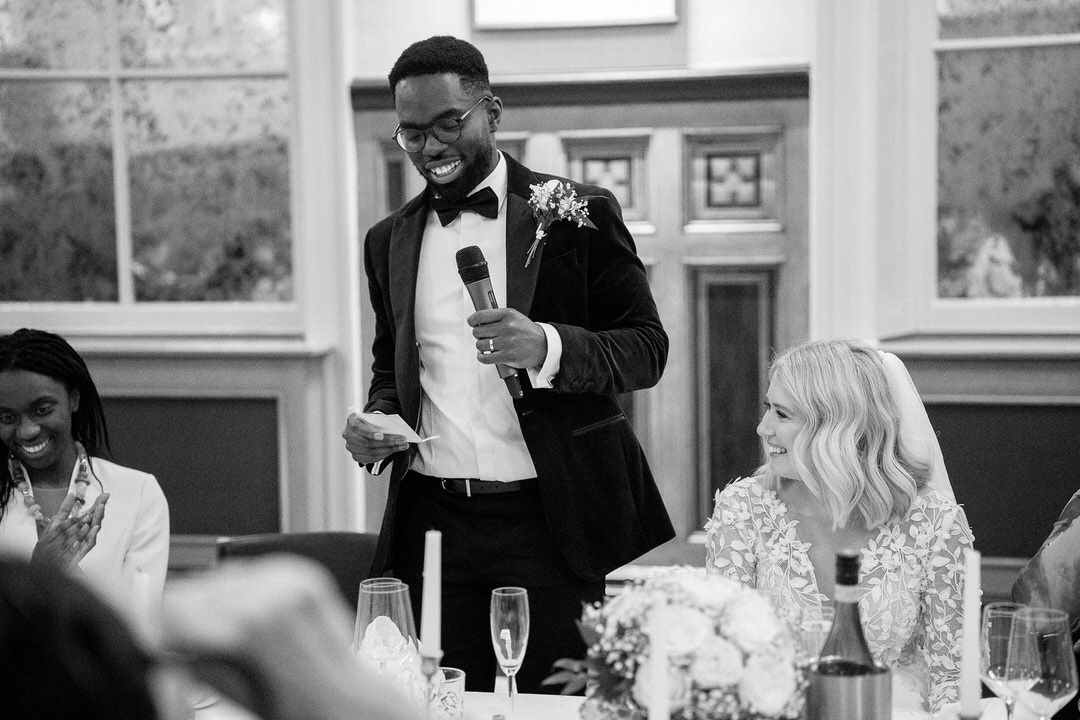 A speaker in a tuxedo and bowtie gives a toast at an elegant wedding reception with floral centerpieces.