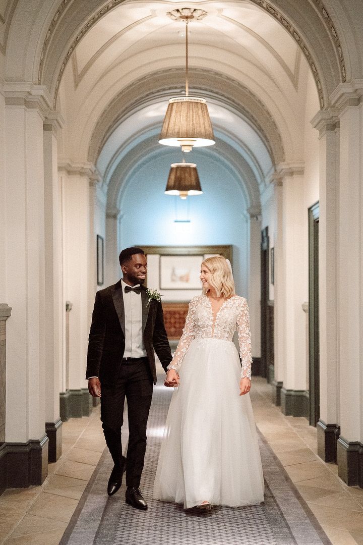 A newlywed couple walking hand in hand down an elegant hallway with arched ceilings and pendant lighting.