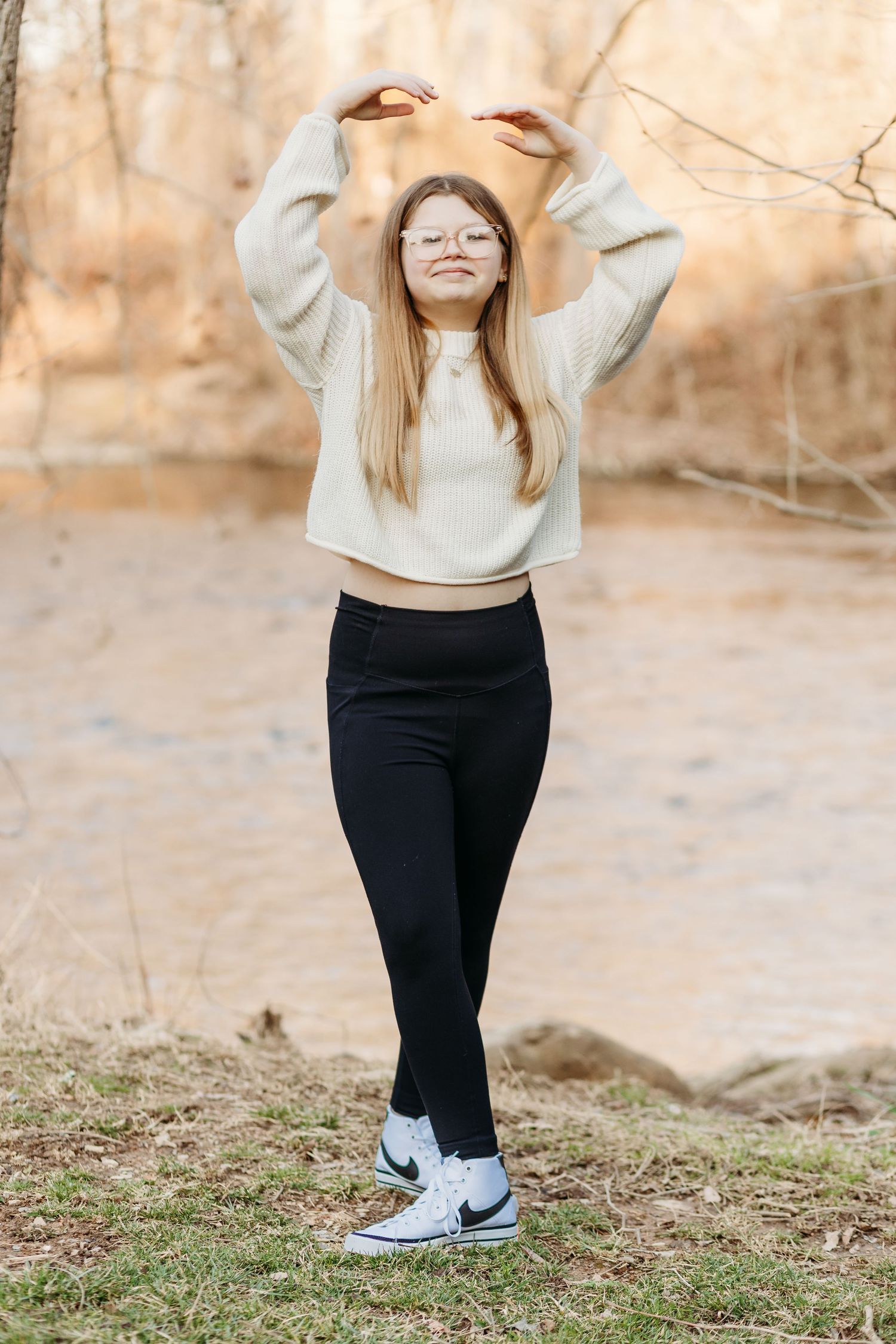 A person wearing a beige sweater and black leggings stands on grass while wearing white Nike sneakers.