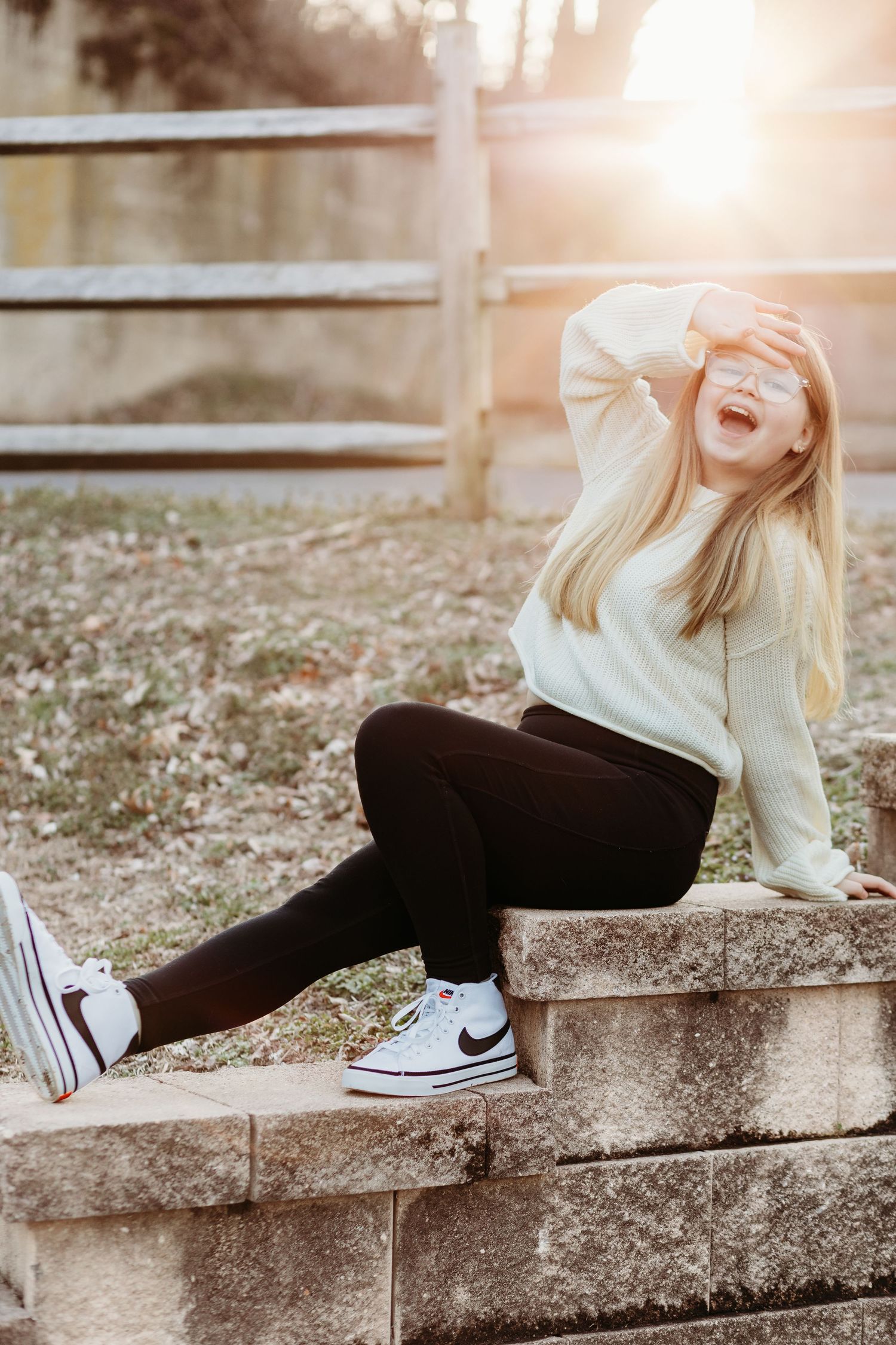 A person in a cream sweater and black pants sits casually on a stone wall during golden hour.