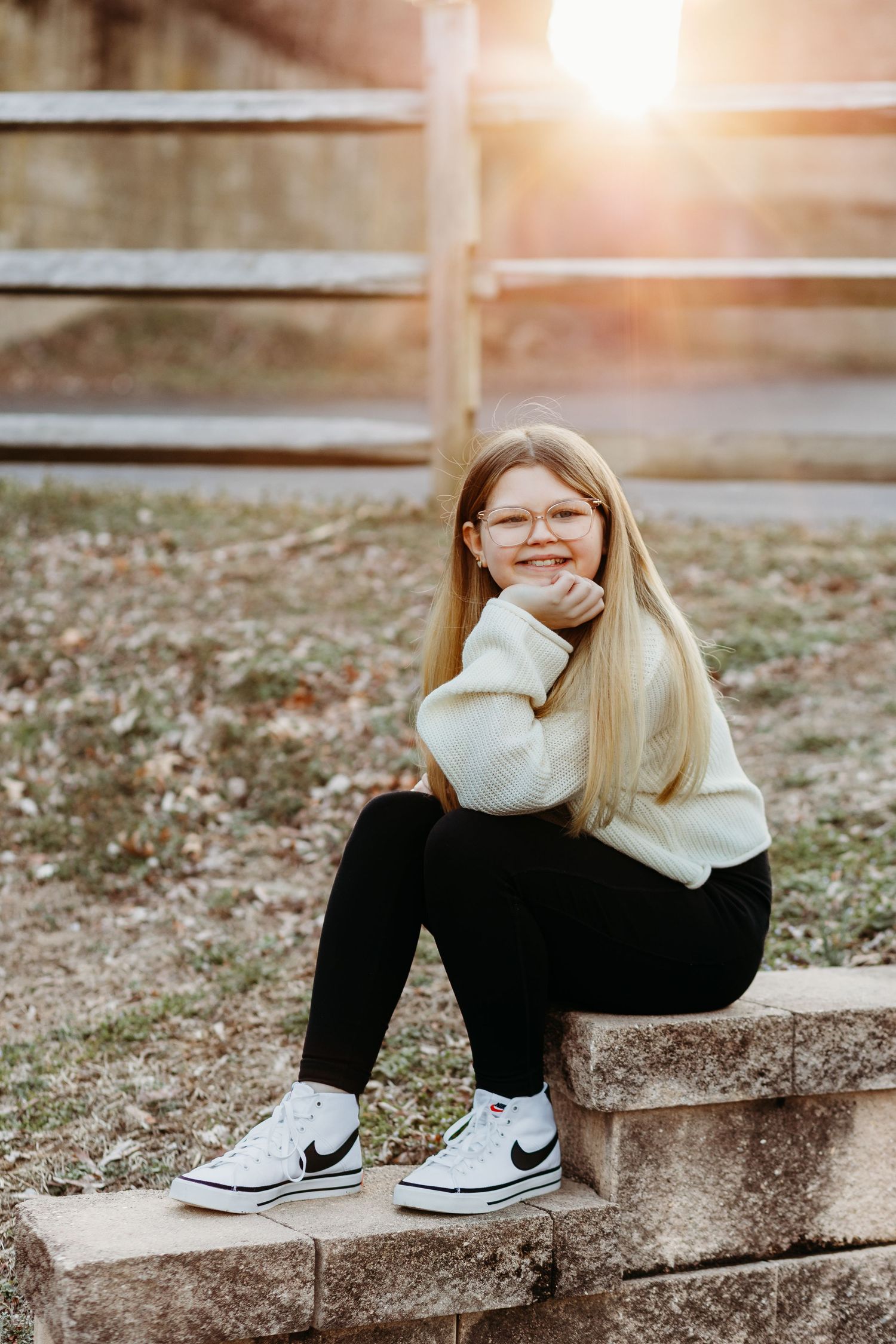 A person in a cream sweater and black pants sits on stone steps at sunset wearing white Converse sneakers.