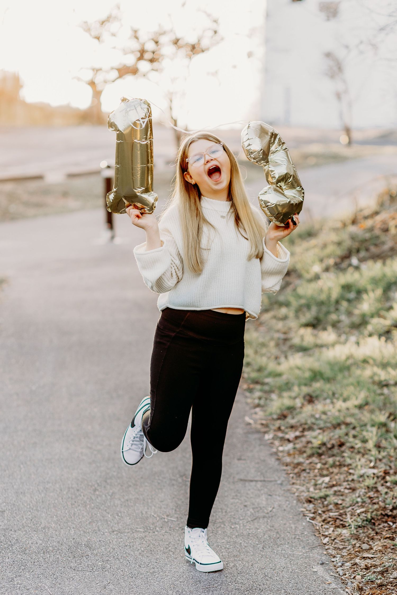 A person wearing a white sweater and black pants jumps playfully on a path while wearing white sneakers.