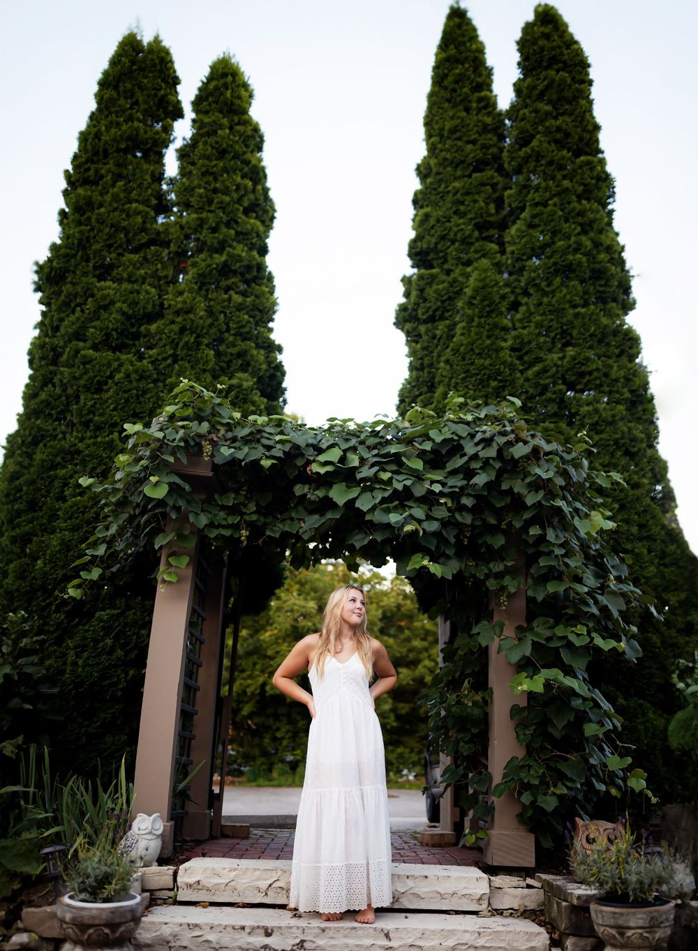 A white sundress is showcased in a garden setting with tall evergreen trees and stone walls creating an elegant backdrop.