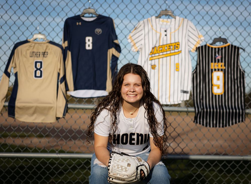 Baseball uniforms and jerseys displayed on chain link fence against blue sky backdrop.