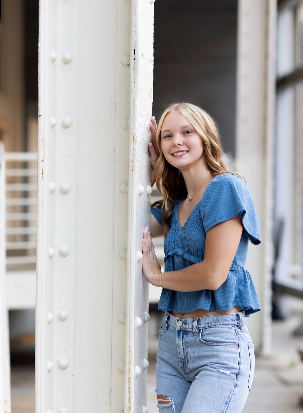 Person in denim outfit poses playfully against white pillars in an outdoor architectural setting.