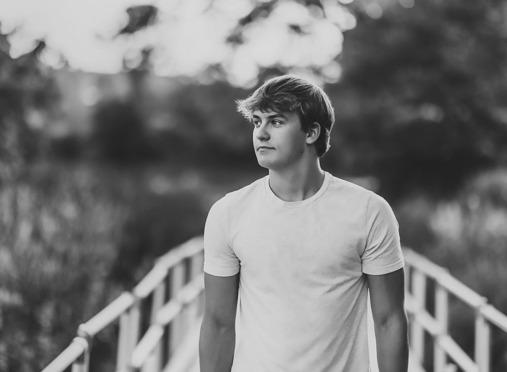 Black and white portrait of someone in a white t-shirt standing on a bridge with a blurred natural background.