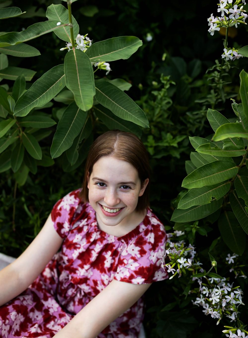 A person in a red and white floral dress poses against a backdrop of lush green foliage and delicate white flowers.
