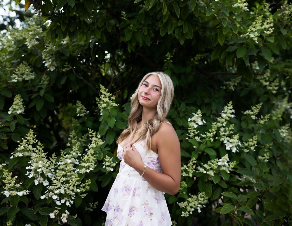 A person in a white floral sundress poses outdoors against lush green foliage on a summer day.