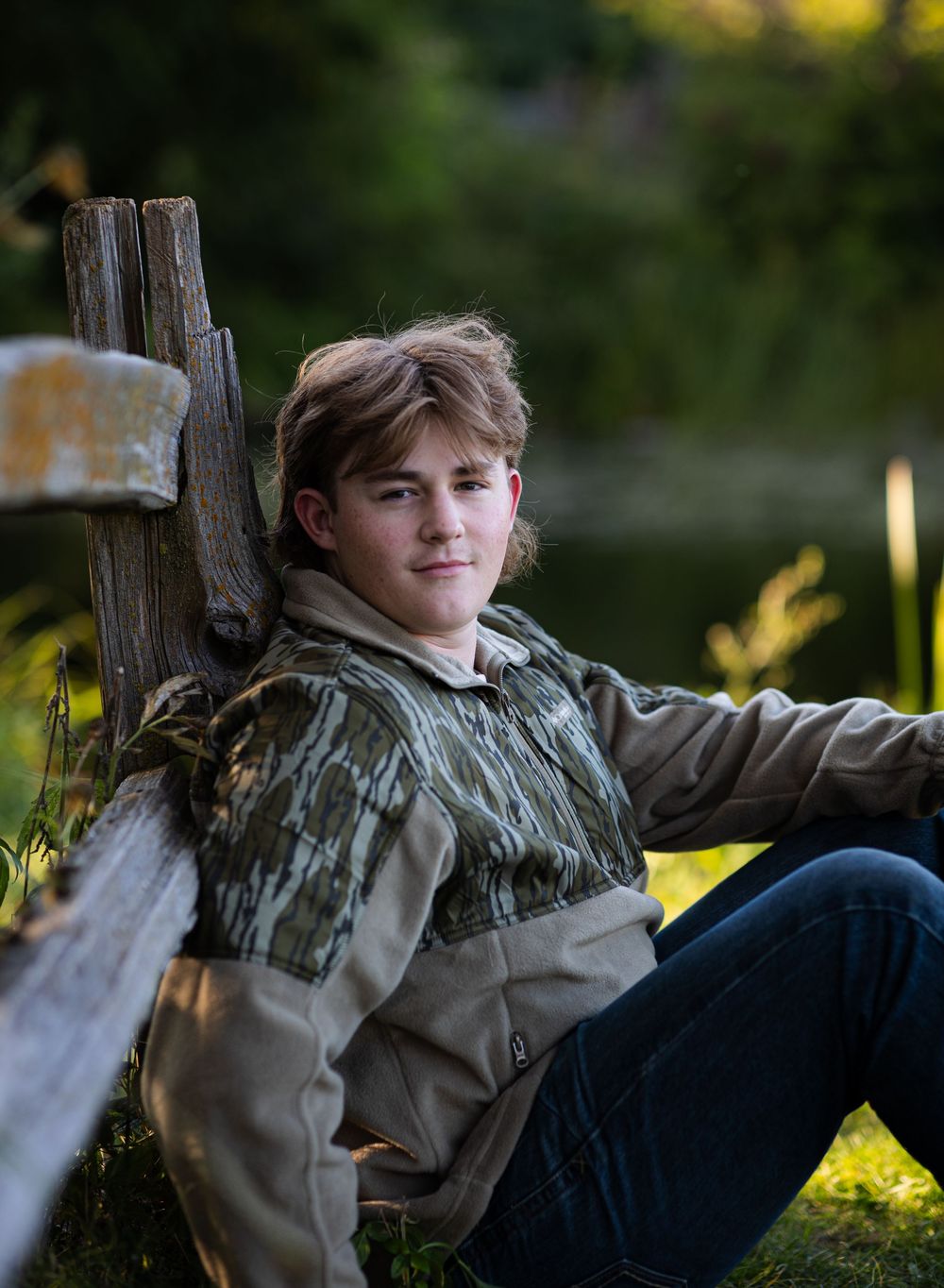 A young person in camouflage clothing relaxes against a rustic wooden fence post in golden afternoon sunlight.
