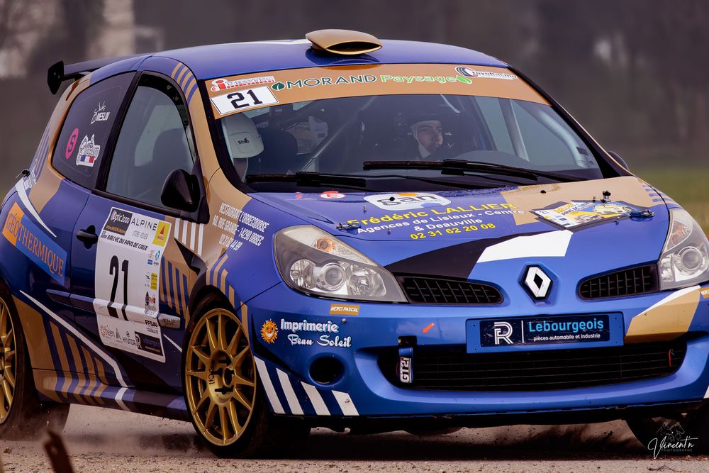 Blue and white Renault Clio R3 rally car racing on a wet asphalt road during competition.