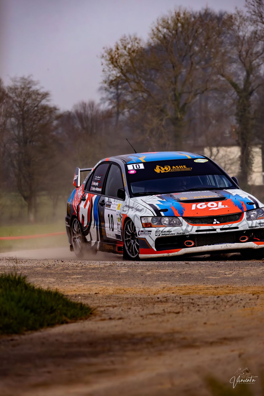 Racing car speeds through dirt track creating dust trail during rally competition on overcast day.