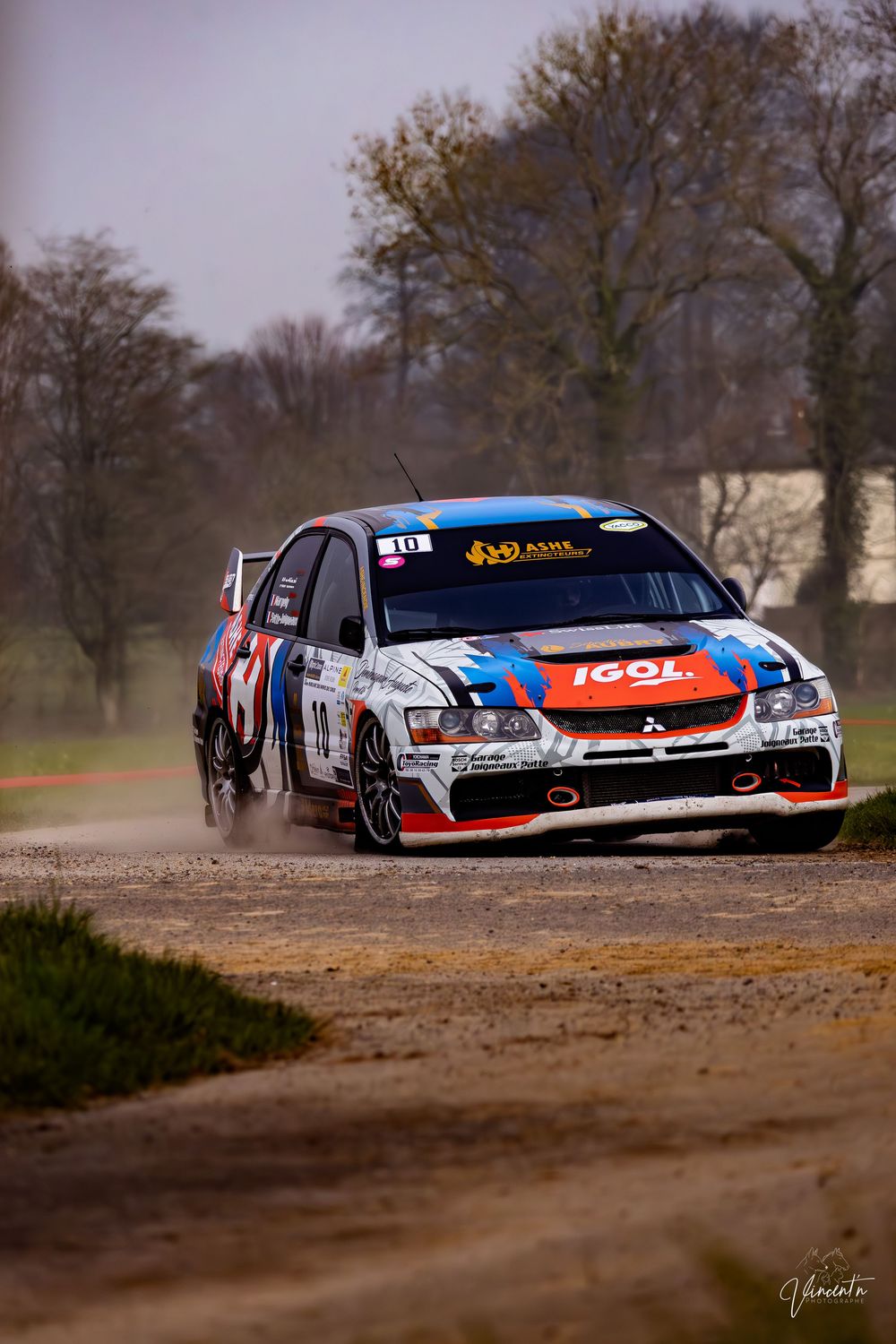 Rally car racing on dirt track with IGOL sponsorship livery kicking up dust during competitive motorsport event.