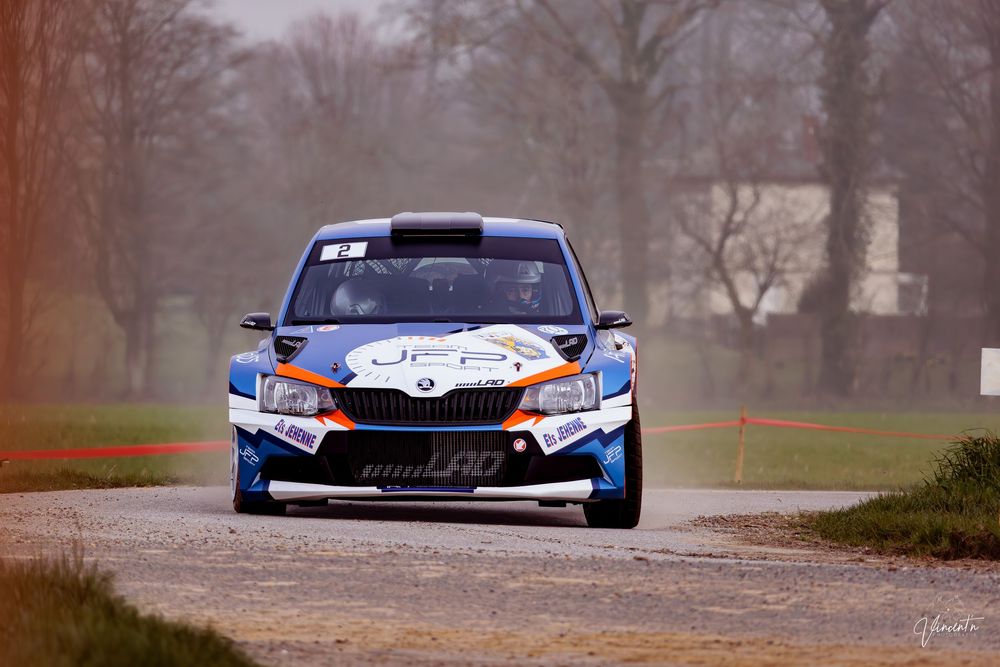 A blue and orange race car navigates a winding track through foggy conditions during a motorsport competition.