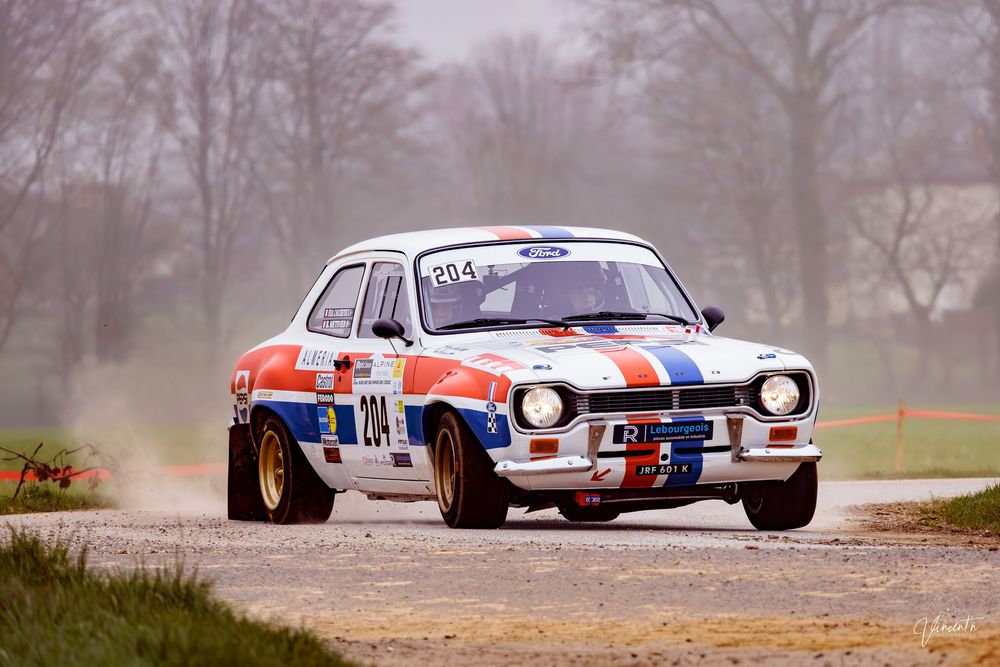 A vintage Ford Escort rally car in red, white and blue livery races on a dusty gravel road during a competition.