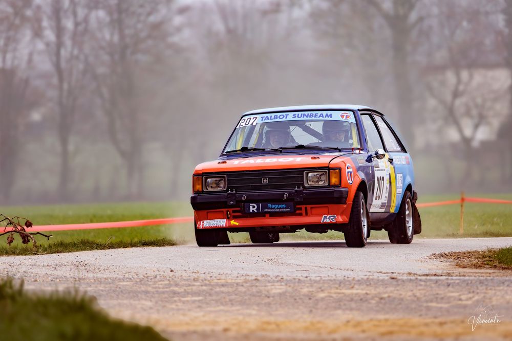 Red and black rally car racing on a dirt track through misty conditions during a competitive motorsport event.