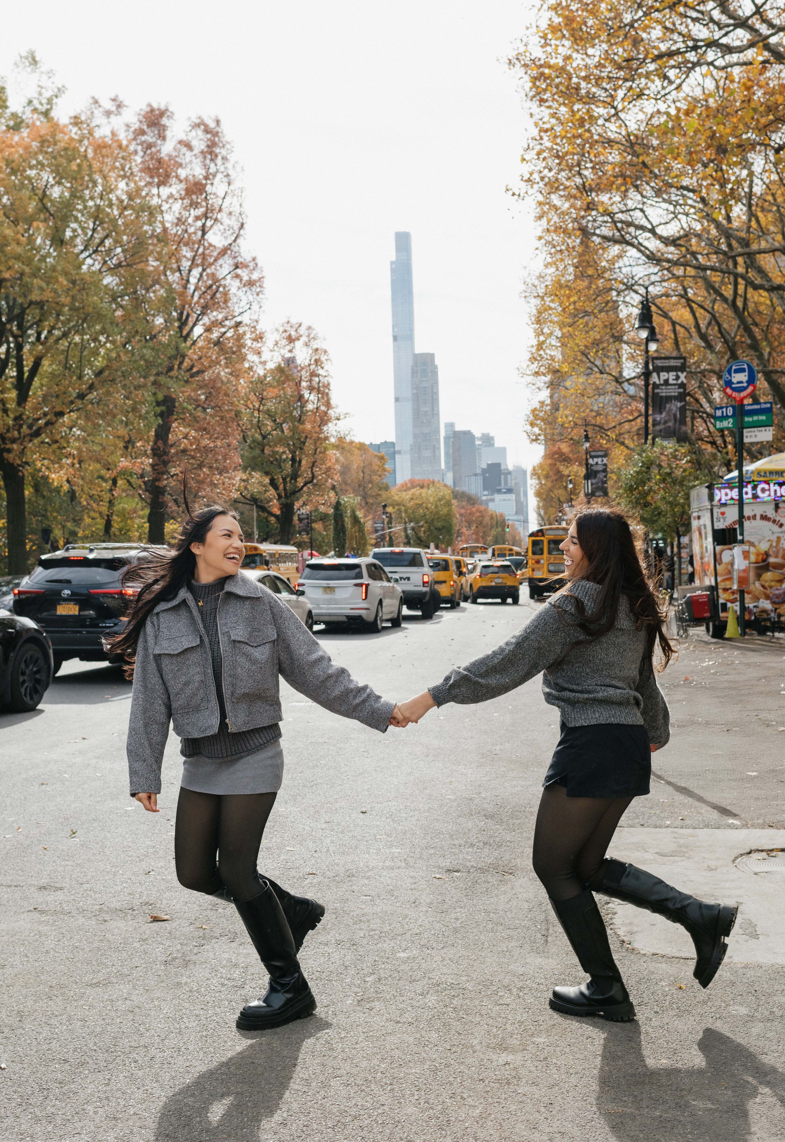 Two people hold hands and dance playfully on a Chicago street with autumn trees and a skyscraper in the background.
