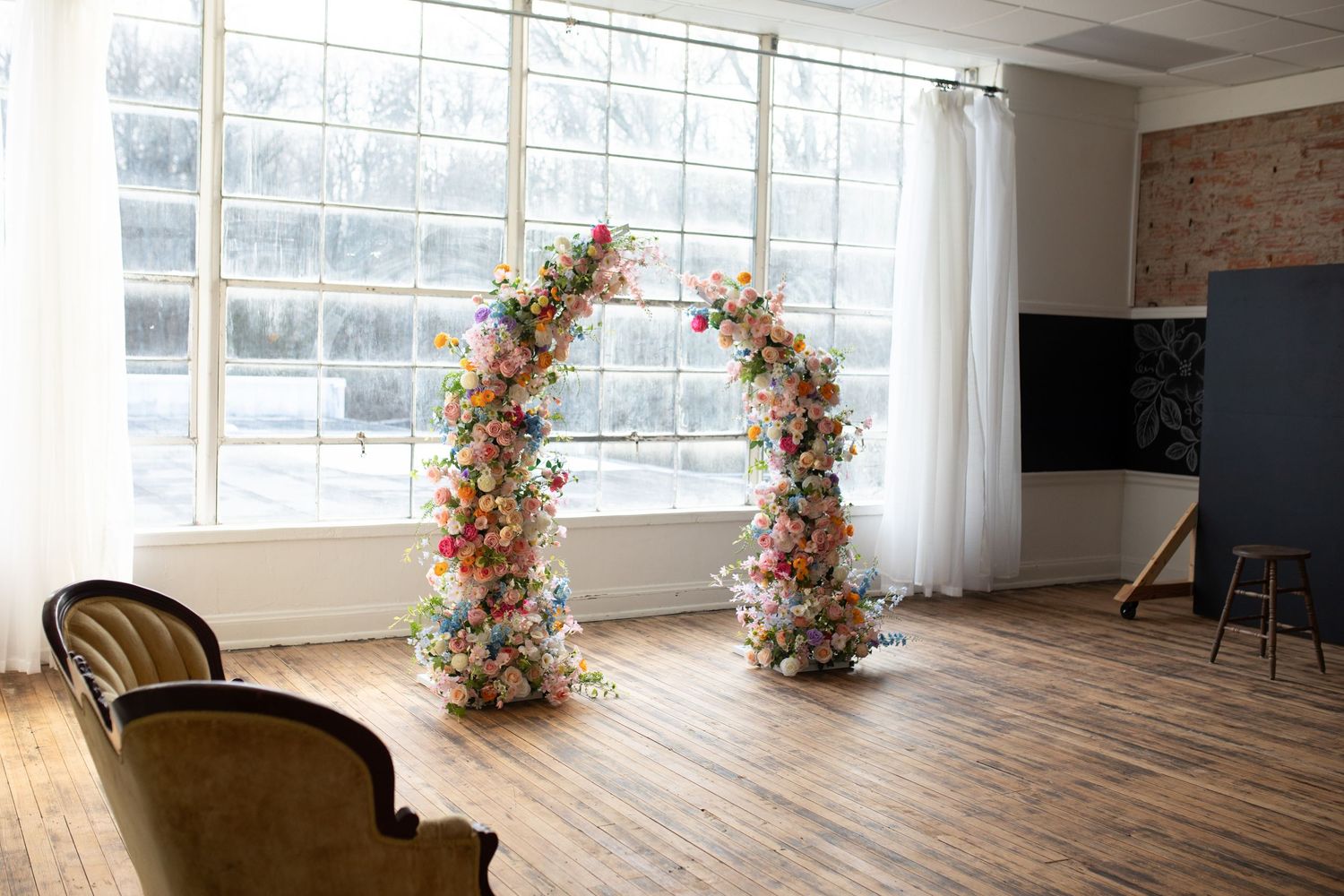 Bright and airy studio space with natural light, hardwood floors, exposed brick wall, and floral arch decorations.