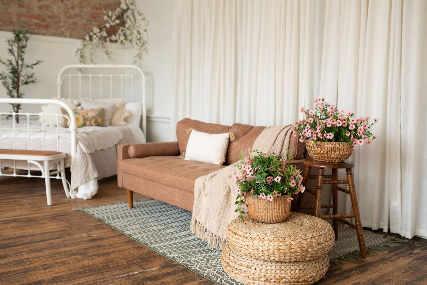 Cozy bedroom interior with pink sofa, white bed, woven poufs, and potted pink flowers on wooden floors.
