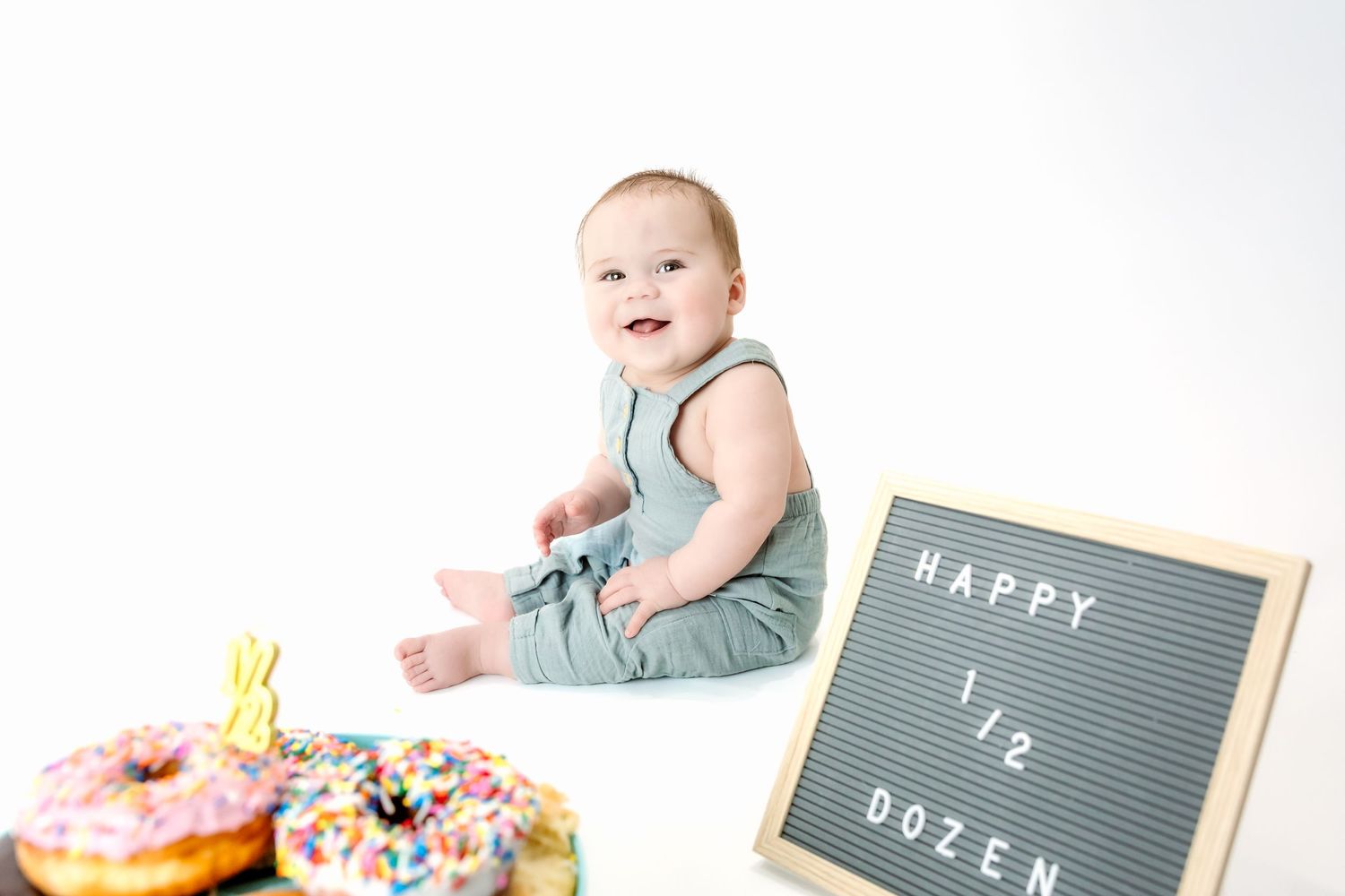 A smiling baby in a gray romper sits beside colorful donuts and a Happy Half Dozen sign.