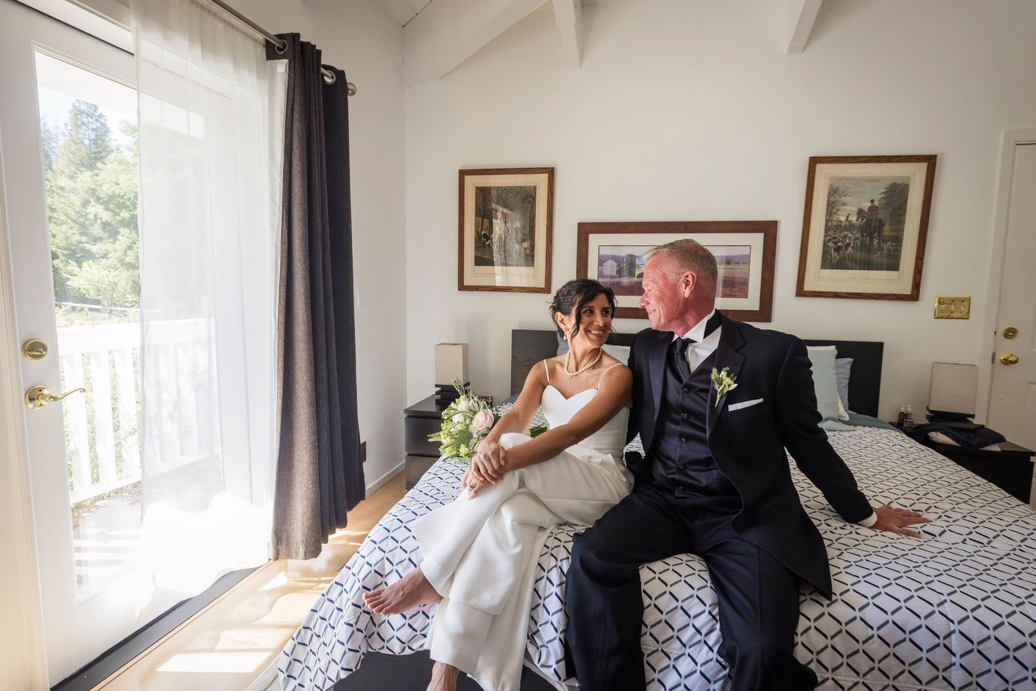 Bride and groom sitting together on a bed in a sunlit room, sharing a tender moment on their wedding day.