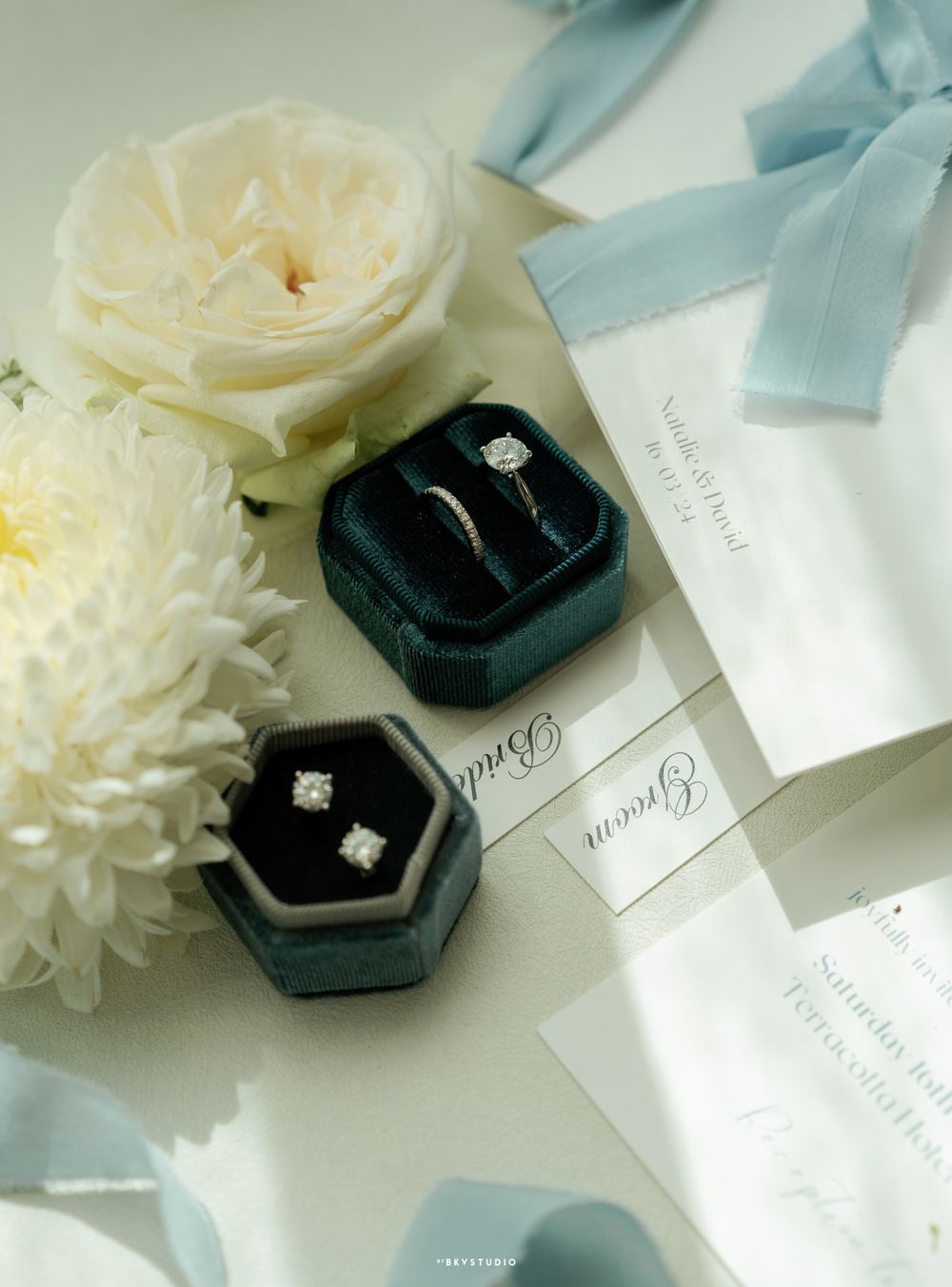 Wedding rings in dark velvet boxes with white roses and blue ribbons on invitations.