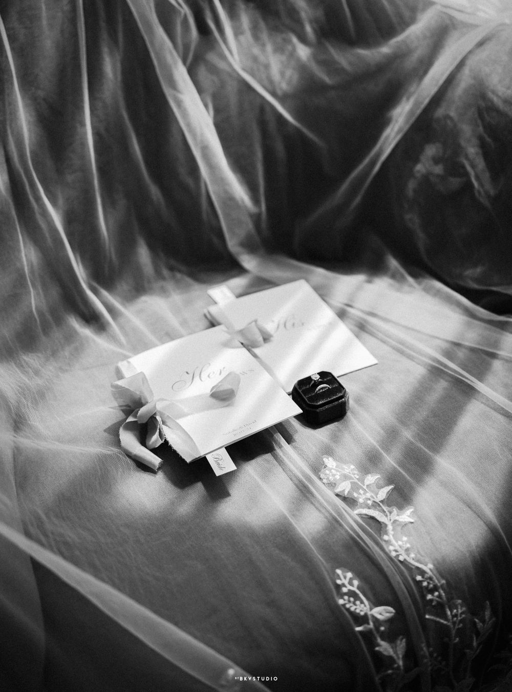 Black and white photo of wedding rings and invitations on flowing fabric.