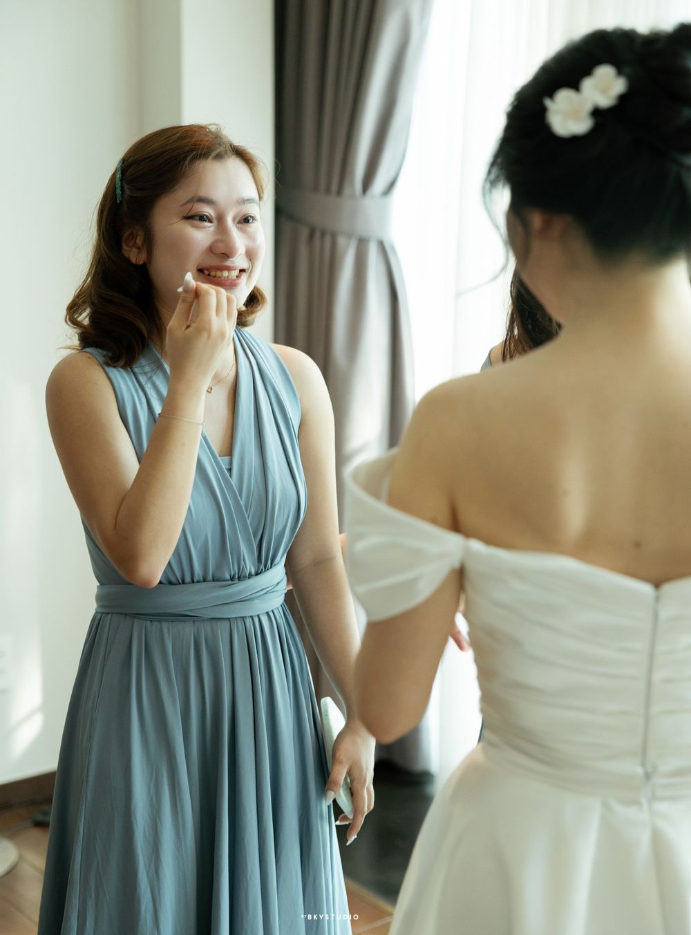 A bridesmaid in a dusty blue dress smiles while admiring her companion in a white wedding gown.