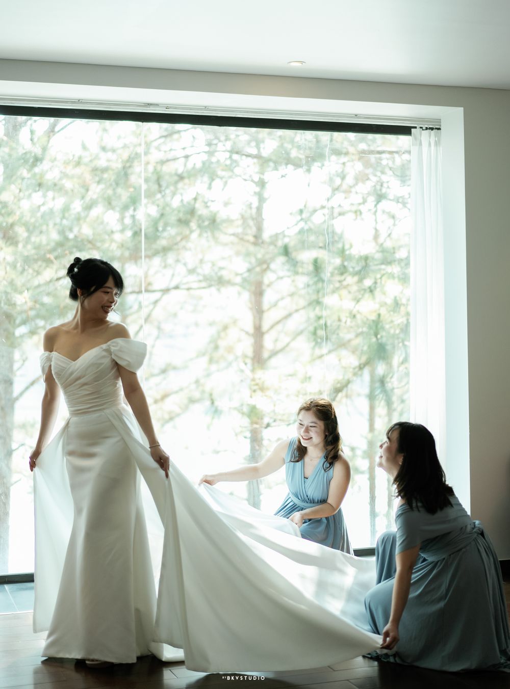 Wedding party helps adjust a flowing white bridal gown near a large window with forest view.