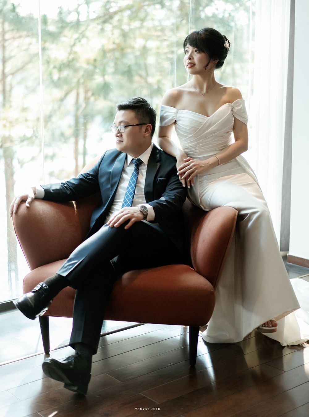Wedding couple poses on brown leather chair against window with forest backdrop.