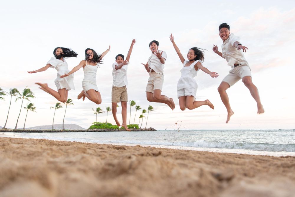 Group of people in white outfits joyfully jumping together on a tropical beach with palm trees and ocean in background of Waialae Beach.
