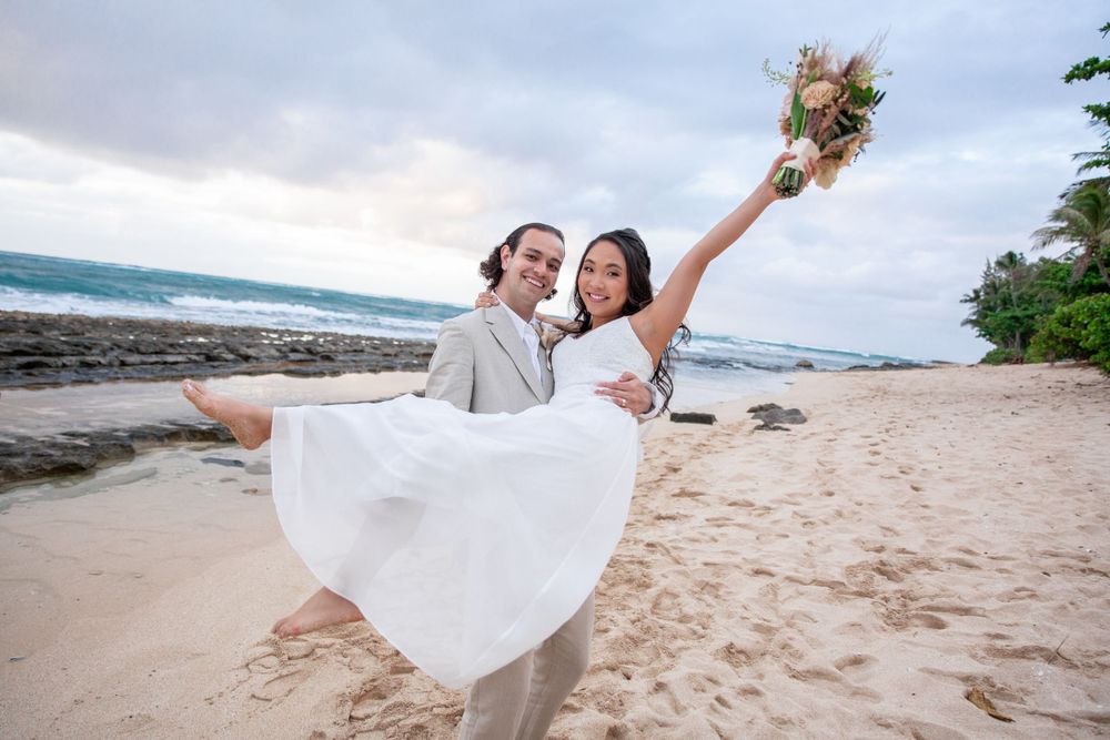 A joyful couple in wedding attire celebrates on scenic Haleiwa beach with crashing waves and palm trees in Hawaii.