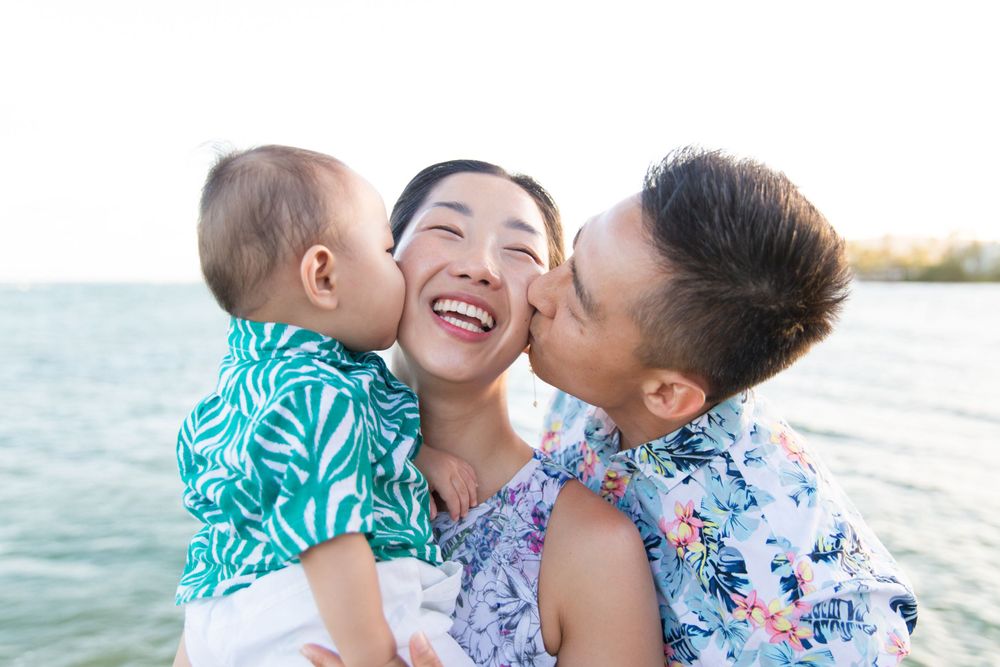 A loving family shares tender kisses and laughter at the beach during sunset with ocean waves in the background.