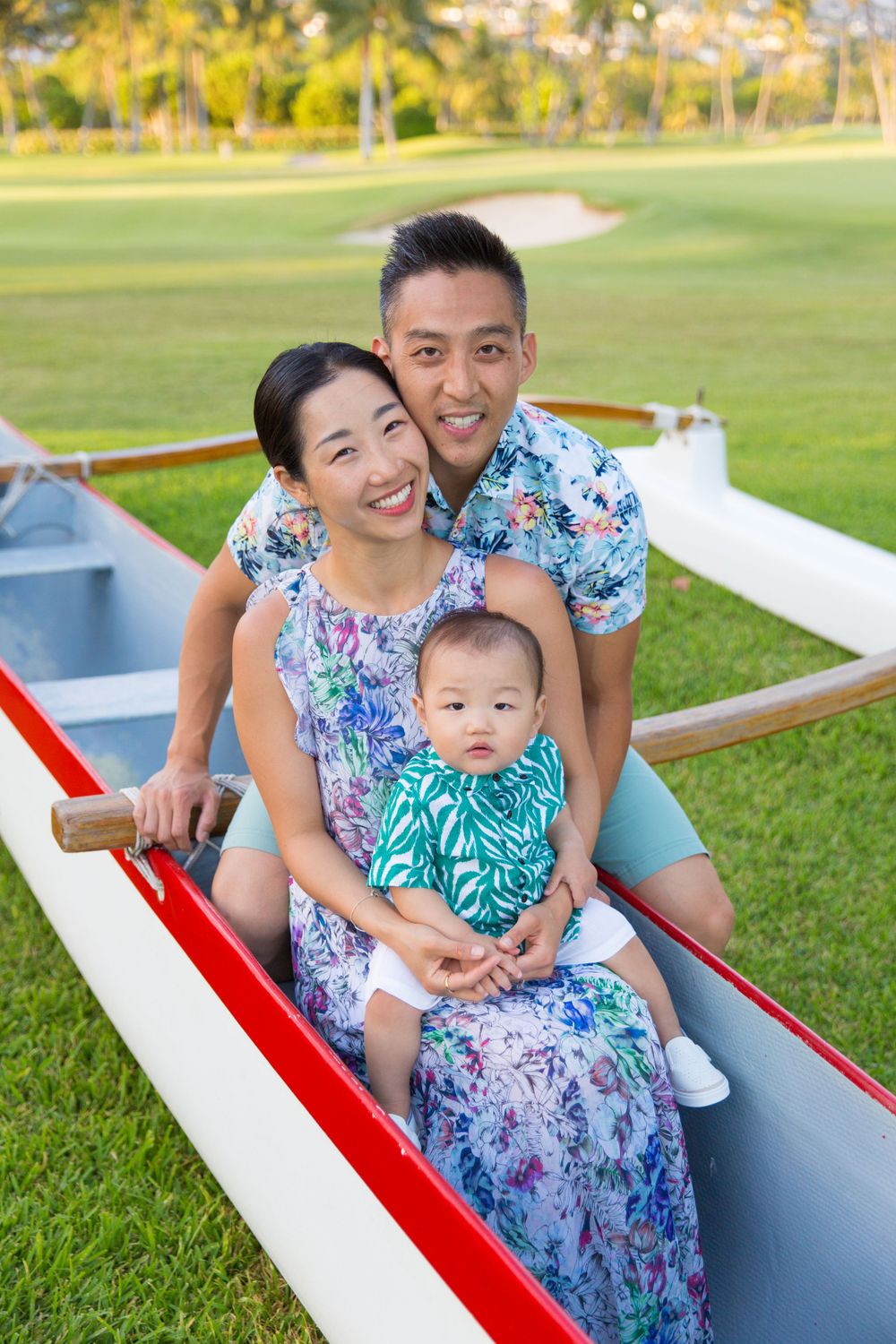 A family wearing floral prints sits together in a white and red outrigger canoe on grass during a sunny day.
