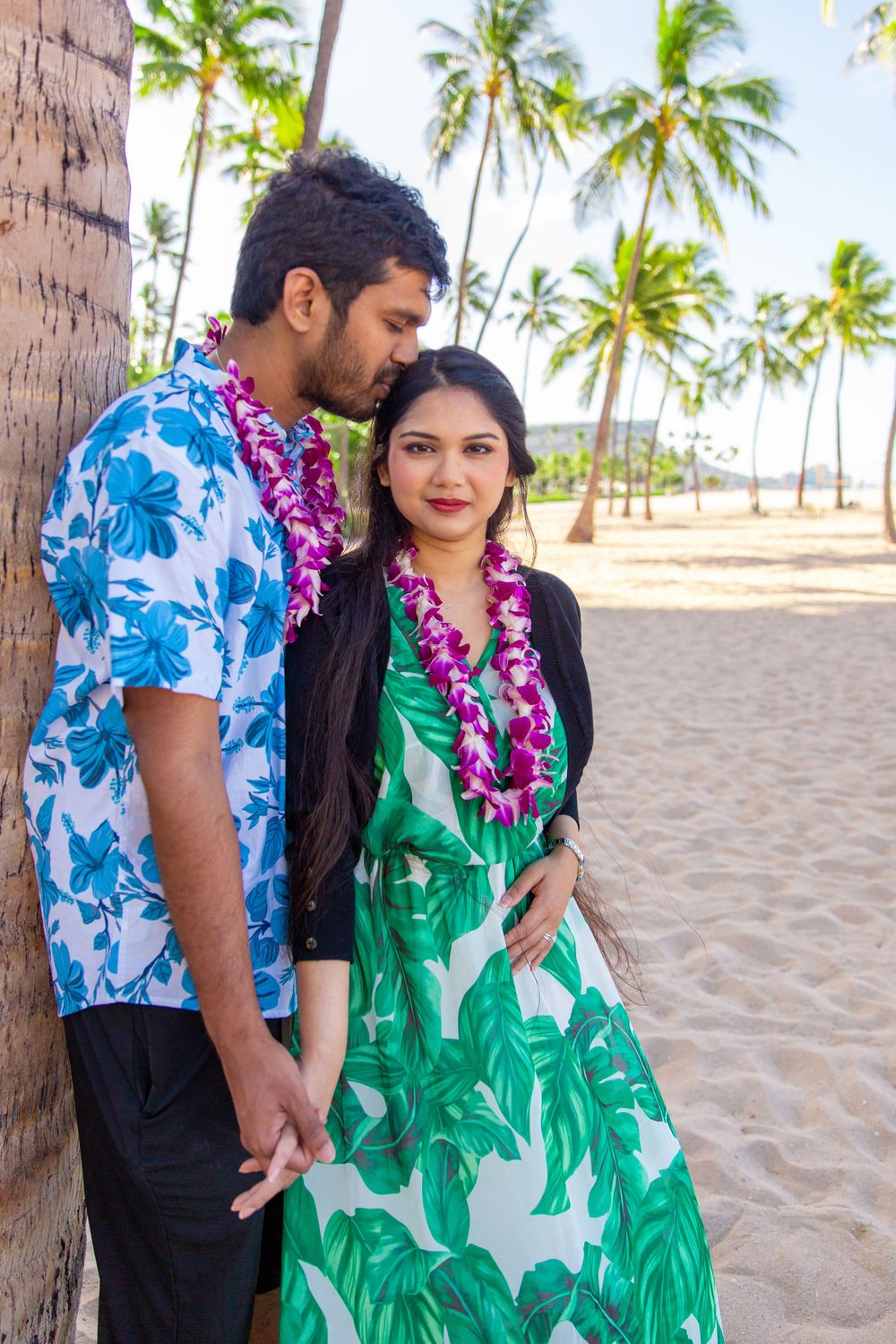 A couple wearing Hawaiian leis and tropical print clothing embrace on a palm-lined beach in Waikiki.