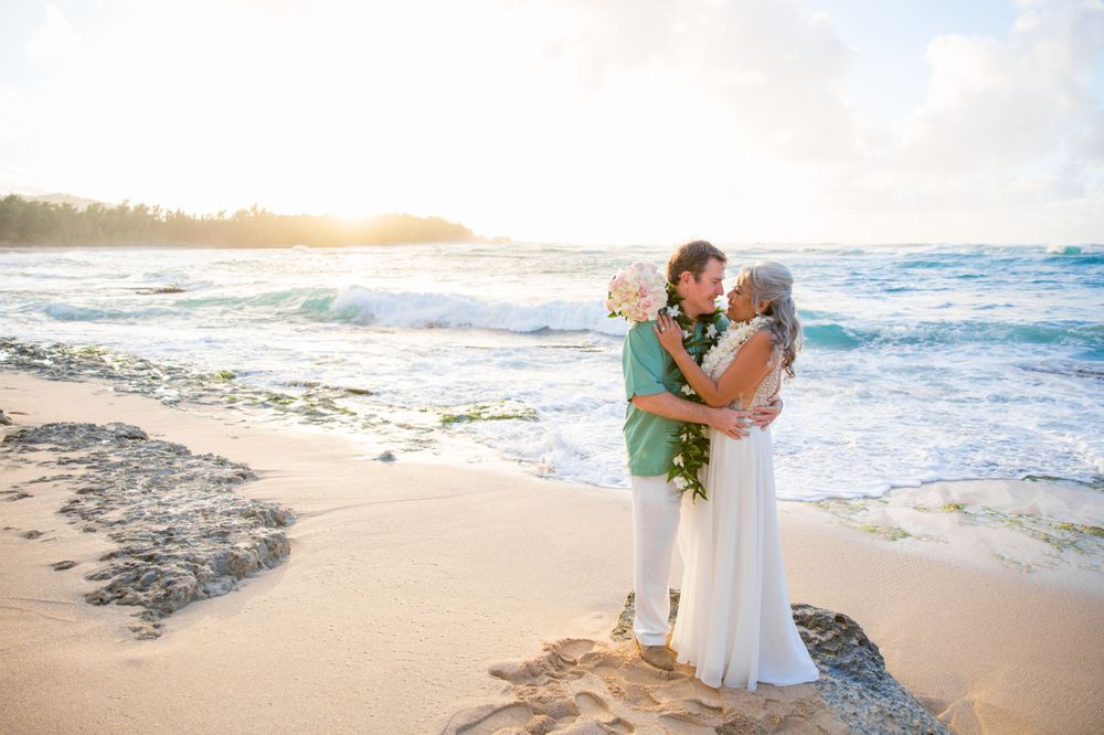 A couple embraces on a sandy beach at sunset with crashing ocean waves and golden sunlight in the background of Turtle Bay.